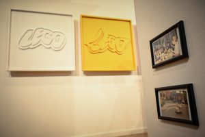 po lewej dwie prace artystyczne przedstawiające przetworzone logo marki Lego, po prawej dwie oprawione fotografie zawieszone na ścianie w galerii sztuki