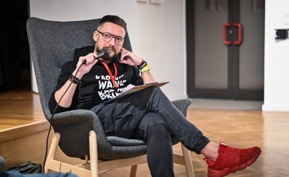 mężczyzna Marcin Zegadło w okularach siedzący w fotelu w bluzie z napisem "kartkowanie rąk nie brudzi" mówiący przez mikrofon