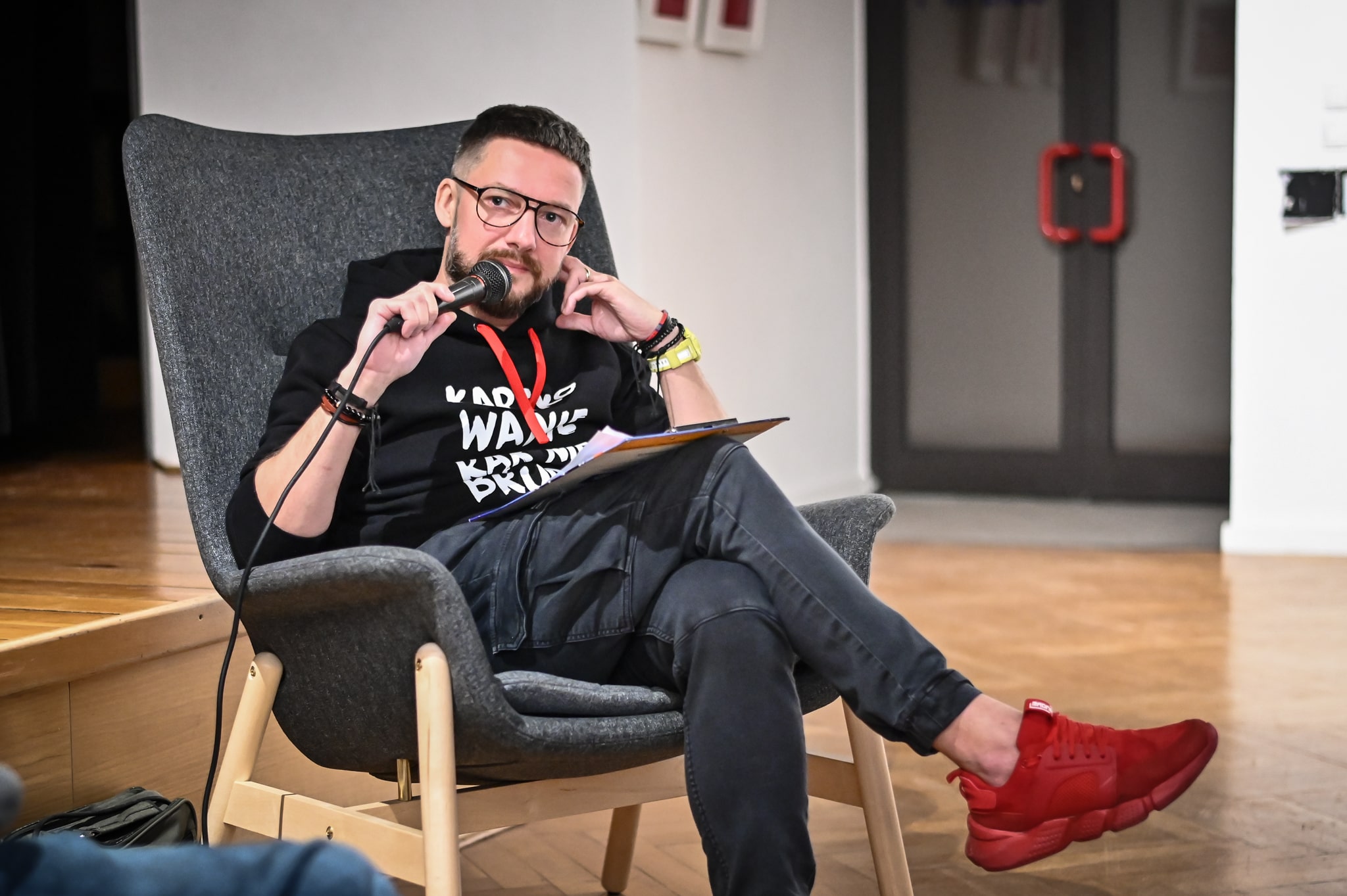 mężczyzna Marcin Zegadło w okularach siedzący w fotelu w bluzie z napisem "kartkowanie rąk nie brudzi" mówiący przez mikrofon