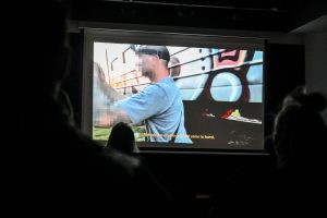 ekran wyświetlający film zatytułowany "Szarańcza", na ekranie mężczyzna z ocenzurowaną twarzą, na dole ekranu żółty napis w języku angielskim: "Nobody has anything but all came to bomb"