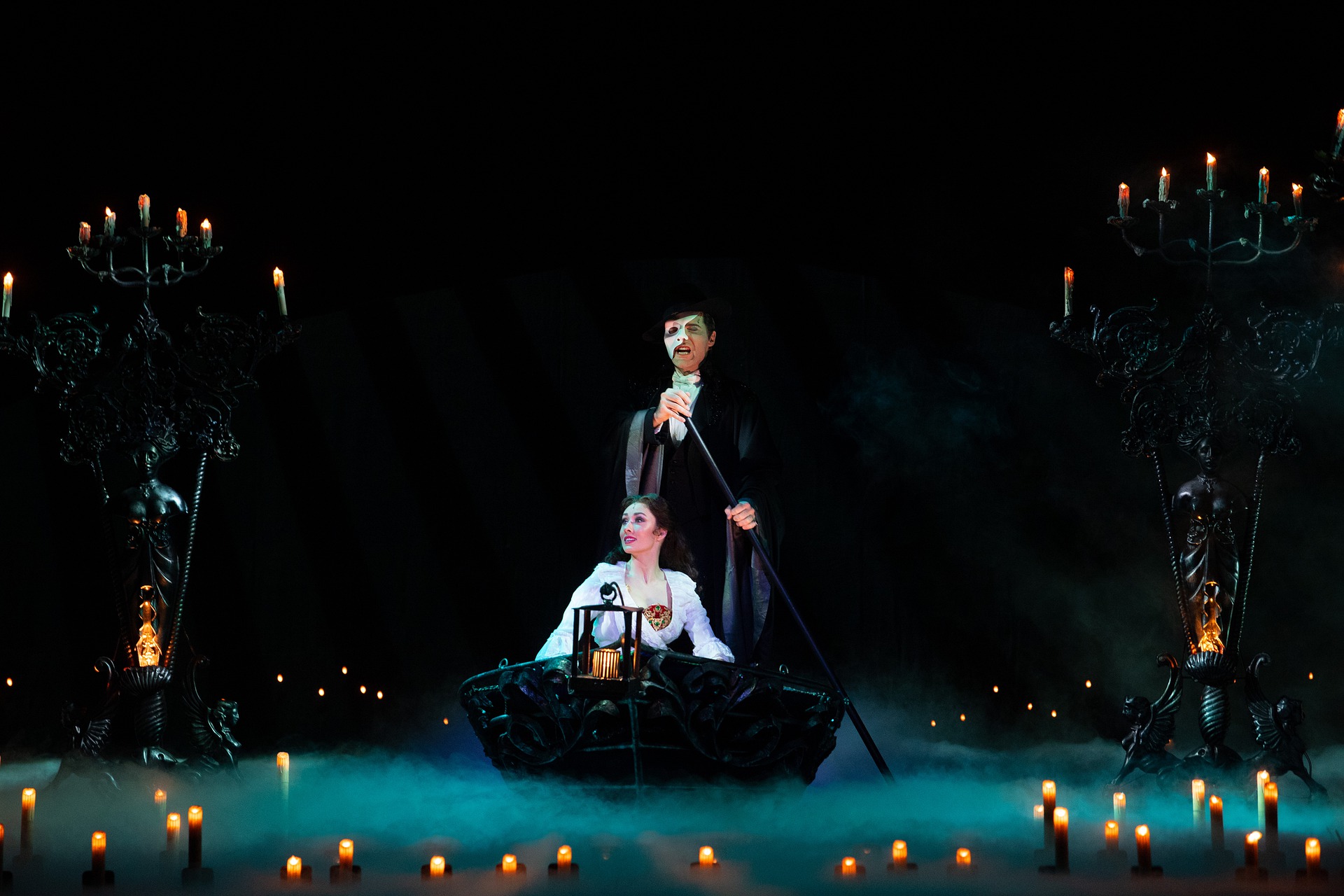 aktorzy operowi na scenie mężczyzna w masce w czarnym stroju wiosłuje stojąc w łódce kobieta w białej sukni siedzi w łódce po obu stronach są świeczniki pełne płonących świec