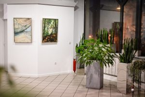 po lewej dwa obrazy malarskie na zawieszone na ścianie w galerii sztuki, po prawej zielone rośliny w wysokich donicach