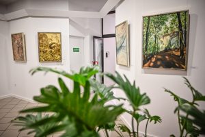 na pierwszym planie liście zielonej rośliny na drugim planie obrazy malarskie zawieszone na ścianie w galerii sztuki