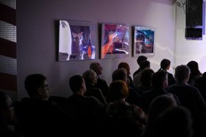 publiczność zgromadzona podczas koncertu a w tle prace plastyczne zawieszone na ścianie obijające występujących na scenie muzyków