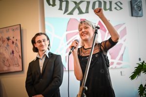 po prawej na pierwszym planie Emilia Dudziec trzymająca mikrofon w prawej dłoni z uniesionym prawym ramieniem na drugim planie po lewej spoglądający na nią Aleksander Szumlas