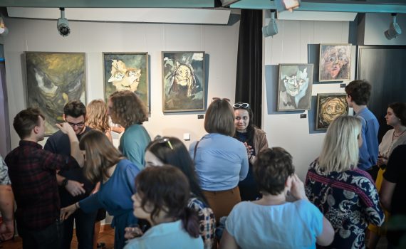 na pierwszym planie publiczność oglądająca obrazy na drugim planie ściana z zawieszonymi na niej obrazami