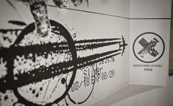 fragment muralu Bartłomieja Stypki przedstawiający samolot pozostawiający za sobą smugę kondensacyjną oraz sygnatura Bartłomieja Stypki w formie znaku X wpisanego w okrąg z napisem zawierającym jego imię i nazwisko oraz poniżej liczba zawierająca aktualny rok 2022