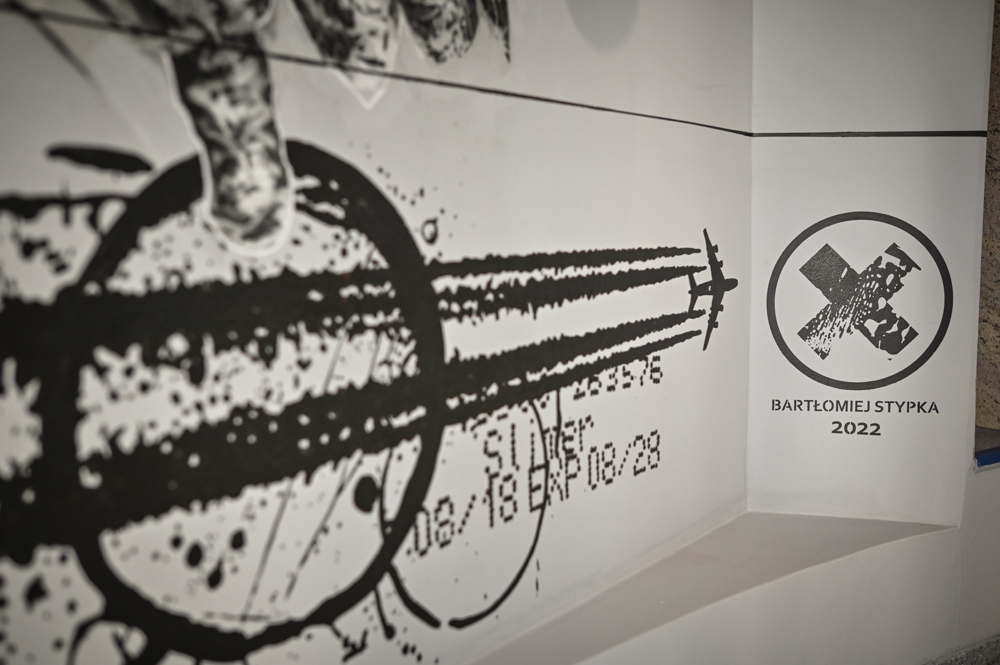 fragment muralu Bartłomieja Stypki przedstawiający samolot pozostawiający za sobą smugę kondensacyjną oraz sygnatura Bartłomieja Stypki w formie znaku X wpisanego w okrąg z napisem zawierającym jego imię i nazwisko oraz poniżej liczba zawierająca aktualny rok 2022